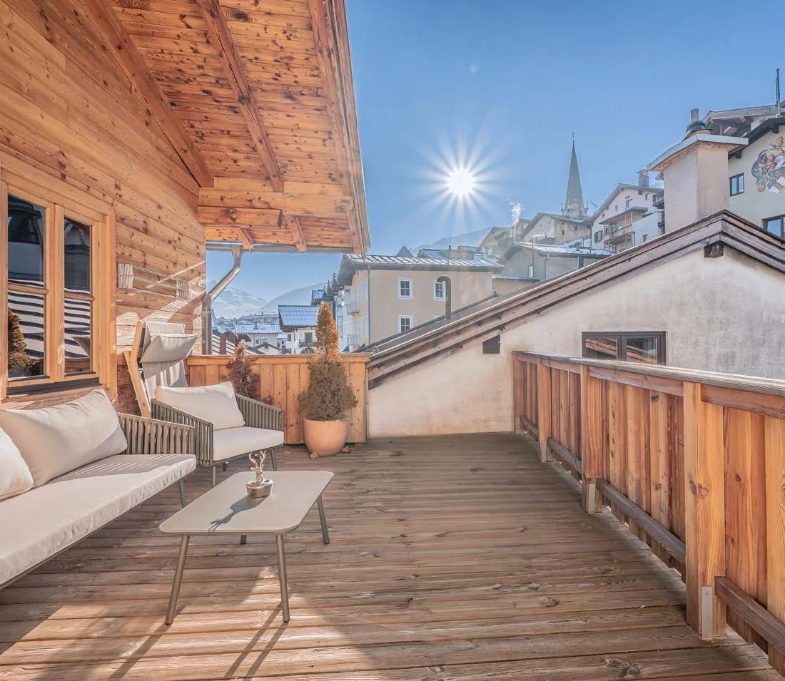 Teilweise überdachte Dachterrasse mit Holzboden, Lounge-Möbeln und Blick auf Kirchturm und Berge in der Sonne