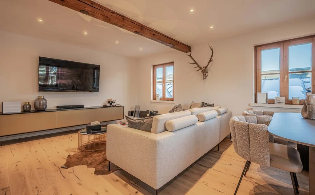 Wohnraum mit alten Holzbalken an der Decke, breiten Holzdielen, heller Sofalandschaft und Fenstern auf zwei Seiten